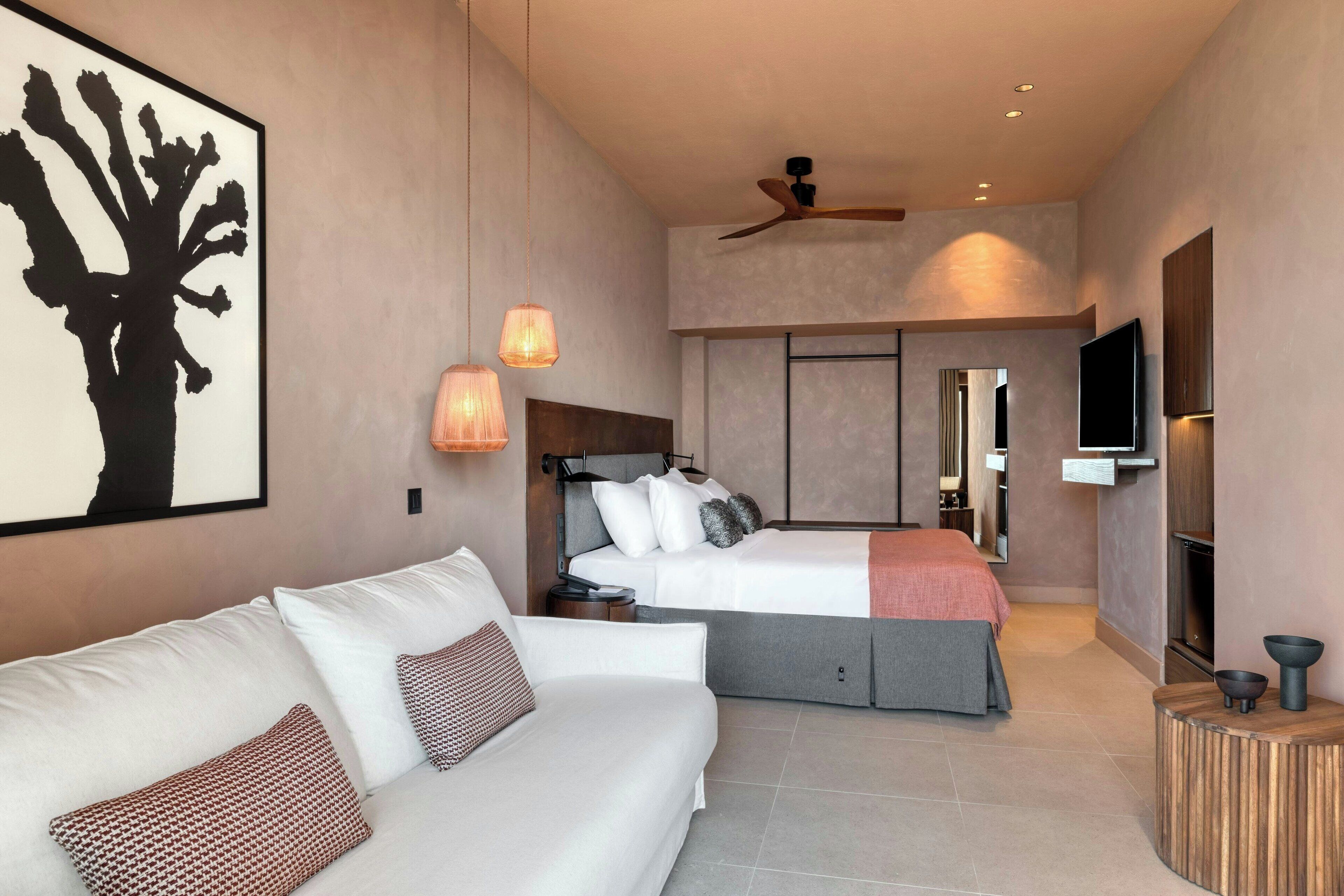 Une chambre d’hôtel moderne avec un canapé blanc, un lit doté d’une tête de lit grise, un ventilateur de plafond, un téléviseur à écran plat et un grand tableau abstrait accroché au mur.