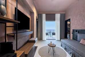 A modern hotel room with a large flat-screen TV, a sofa, a small round table, and a balcony with a view. - Domes of Corfu Autograph Collection (Corfu)