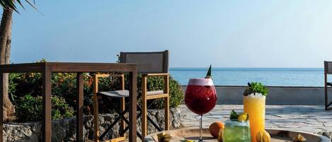 A wooden table with drinks and snacks on a beach with chairs and a palm tree.