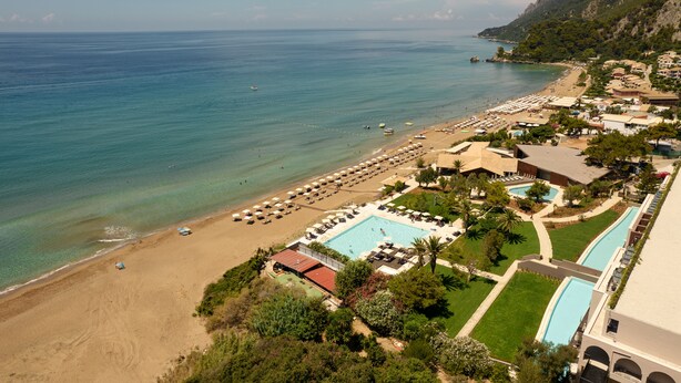 Vista aérea de un complejo turístico costero con playa, piscina y montañas al fondo.