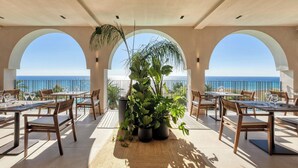 Un espacio de comedor al aire libre con mesas y sillas, plantas en macetas y vistas al mar.