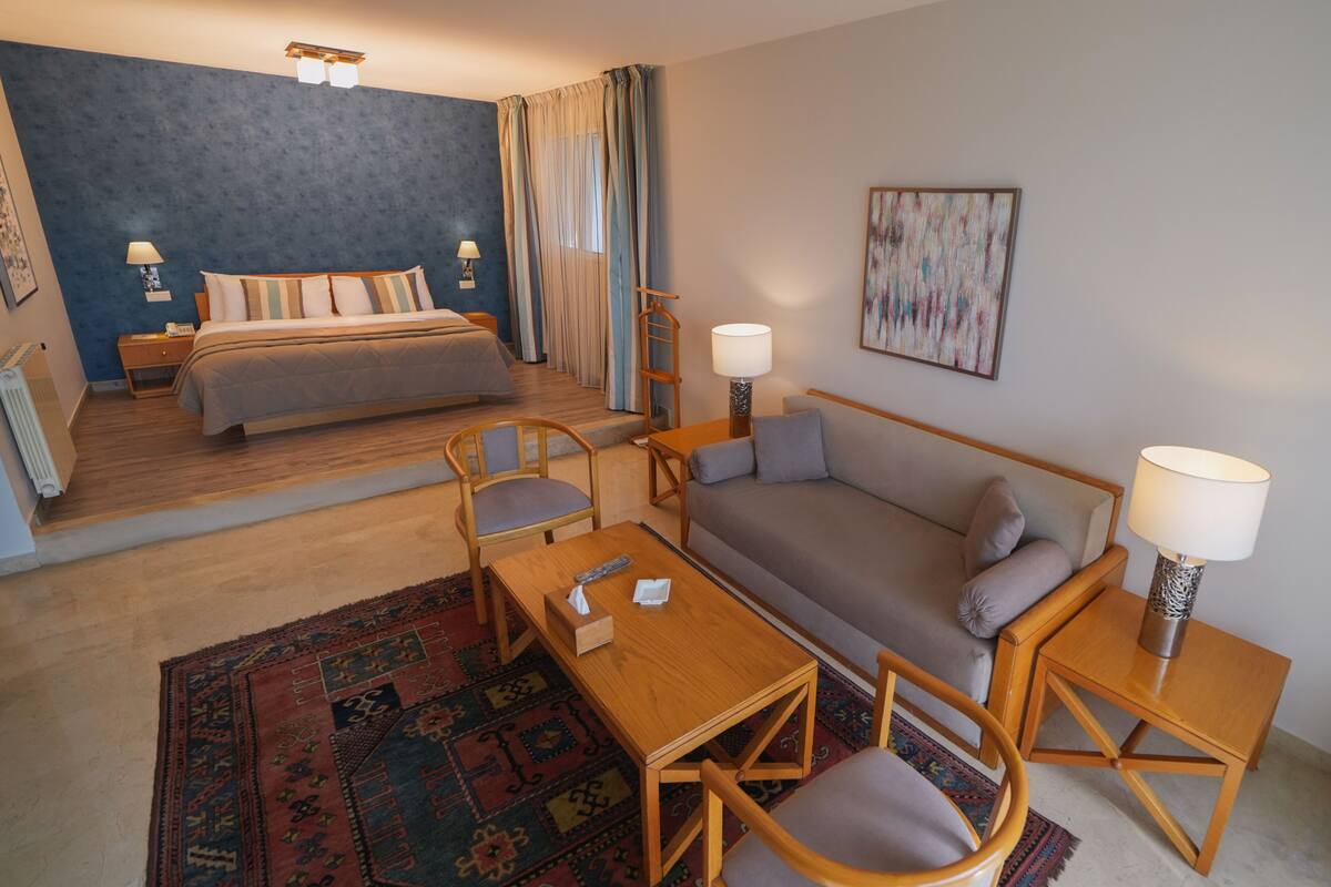 A hotel room with a bed, a sofa, a coffee table, and a chair. There is a window with curtains and a painting on the wall.