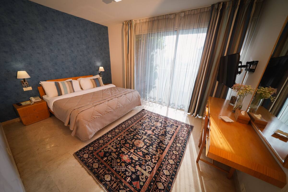 A hotel room with a bed, bedside lamps, a wooden desk, a mirror, and a large window with curtains.