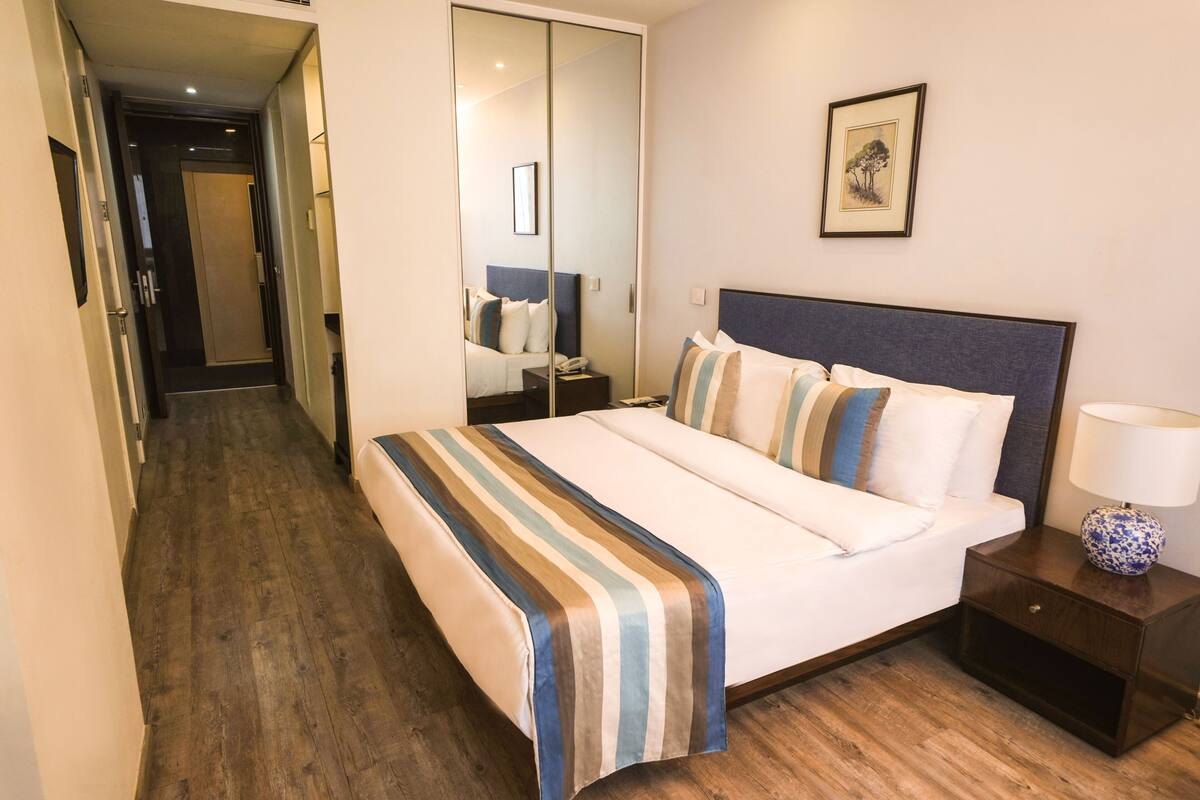 A hotel room with a large bed, a bedside table with a lamp, a nightstand, a wardrobe, and a mirror.