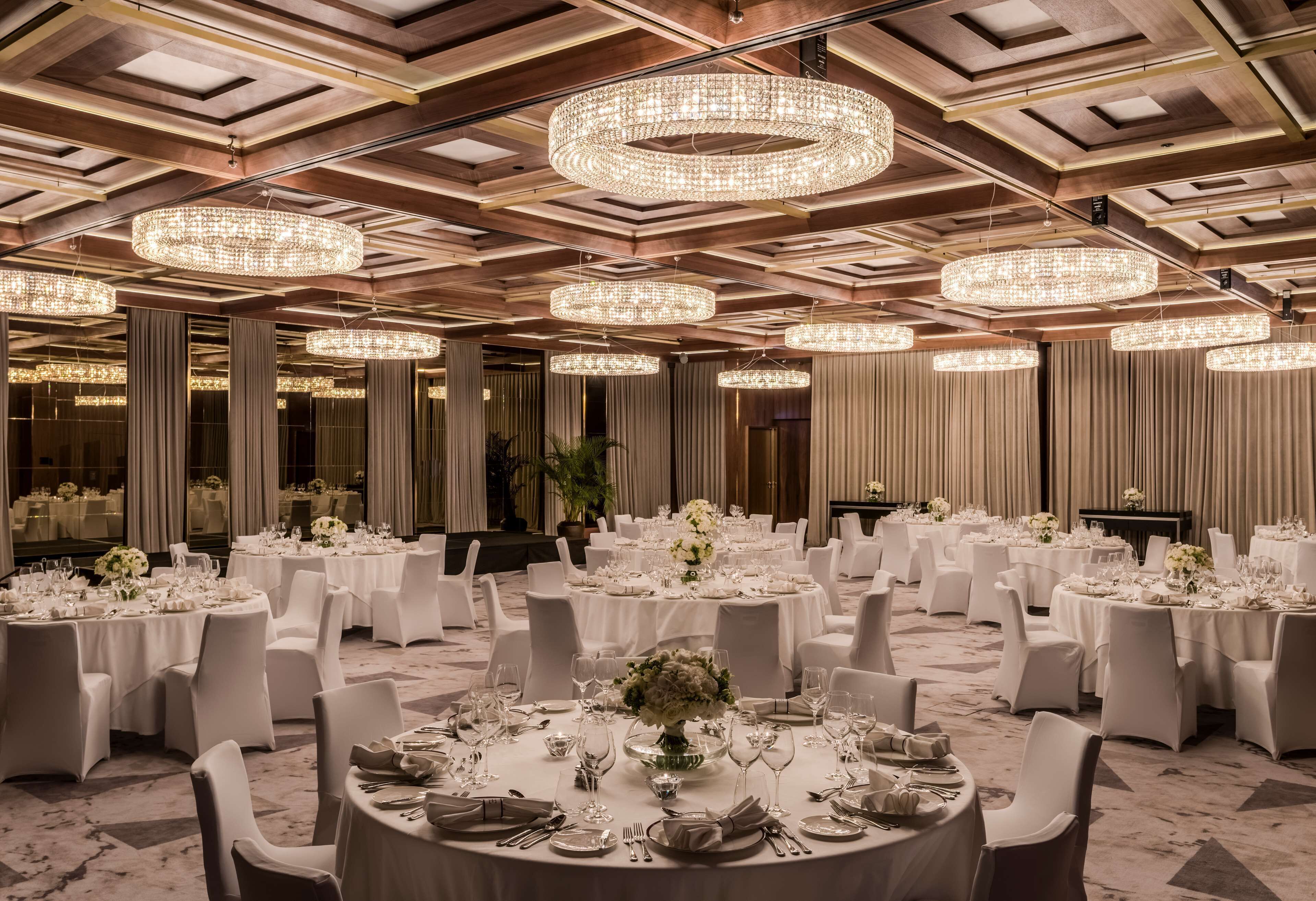A banquet hall with round tables set for dining, large chandeliers, and a high ceiling with wooden beams.