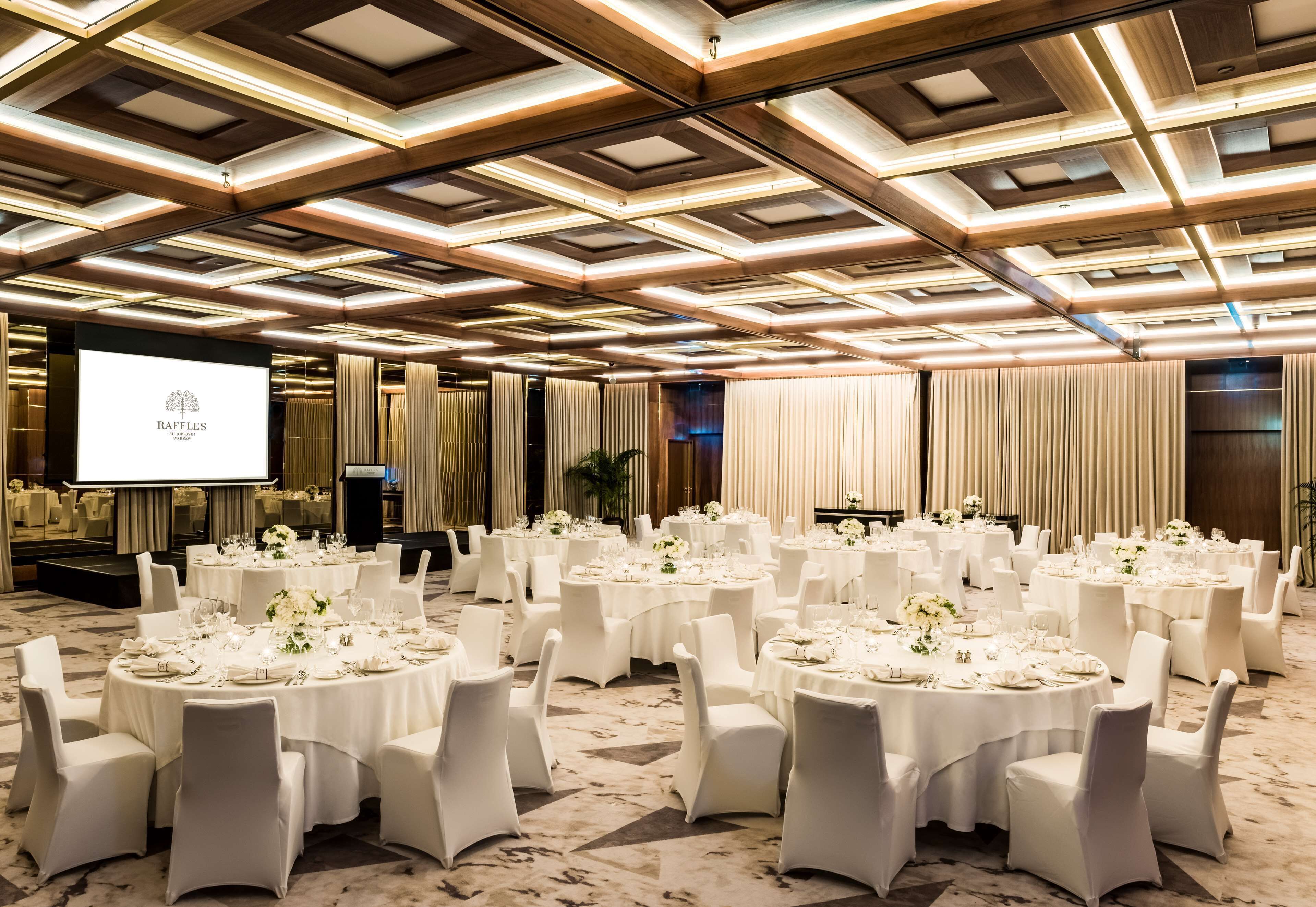 Una sala de banquetes amb taules rodones preparades per a esdeveniments, amb un disseny de sostre modern i una pantalla de projecció que mostra el nom "RAFFLES".