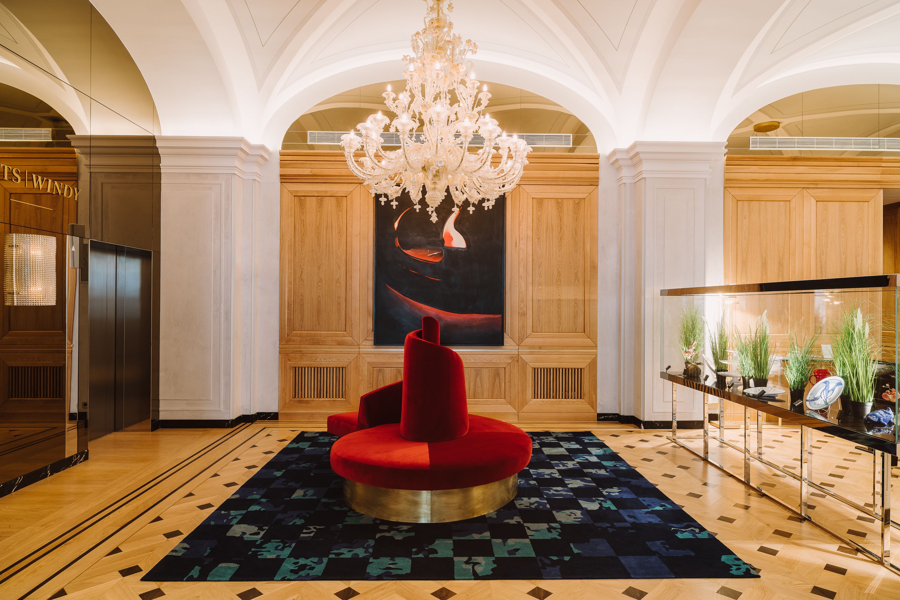 A lobby with a chandelier, a red circular seating area, a large abstract painting, and a plant display.