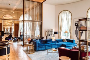 A spacious living area with a large blue sofa, a red armchair, and a glass coffee table. There is a bookshelf with books and a lamp on a side table.