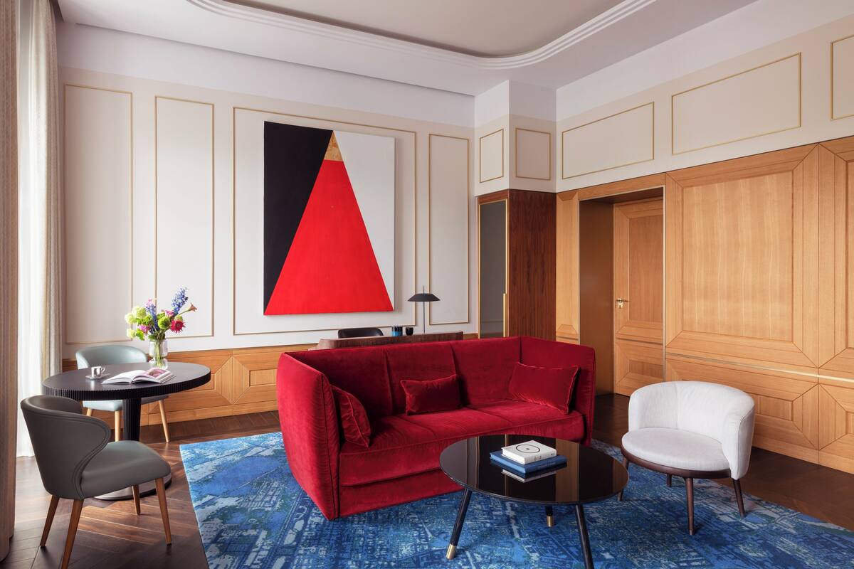 A modern hotel room with a red sofa, a white armchair, a round coffee table, and a blue patterned rug.
