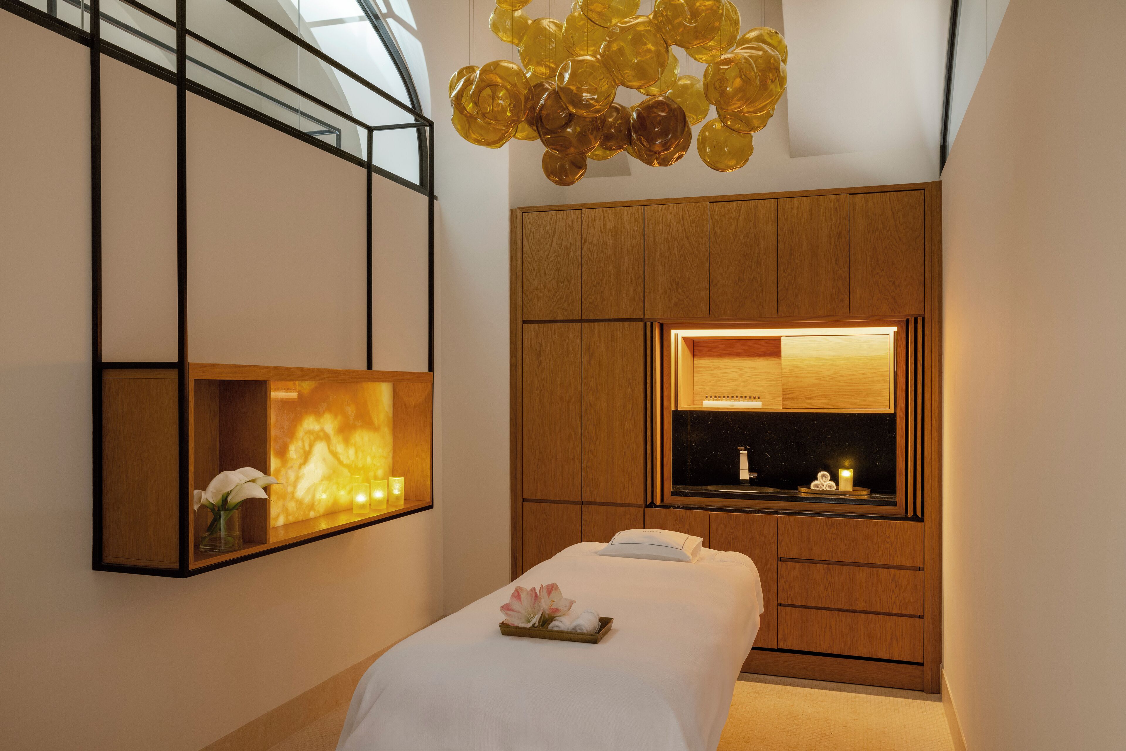 A modern hotel room with a bed, a wooden wardrobe, a mirror, and a unique ceiling light fixture.