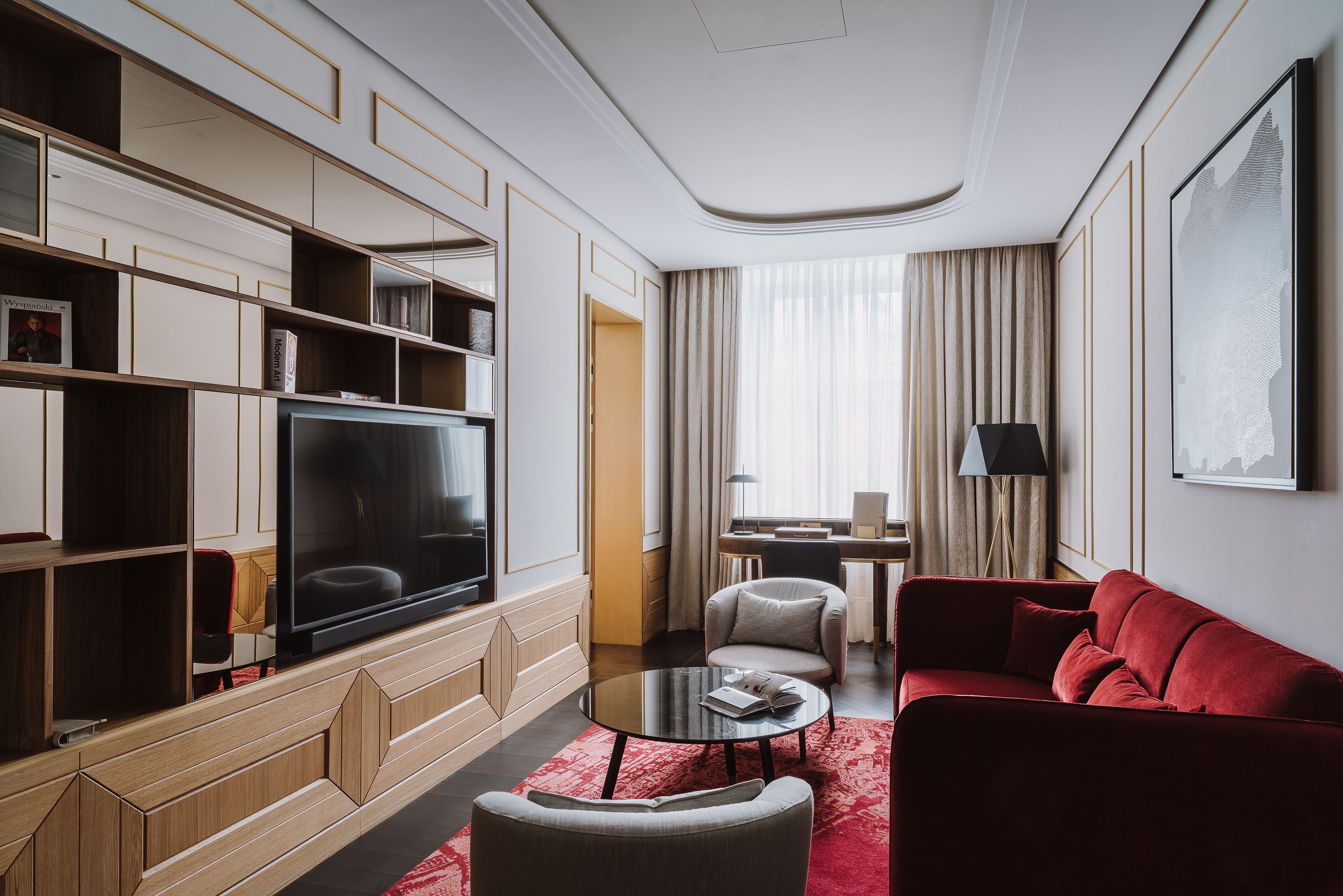 Una habitación de hotel moderna con una televisión de pantalla plana, un sofá rojo, una mesita de centro redonda, una estantería y un escritorio con lámpara.