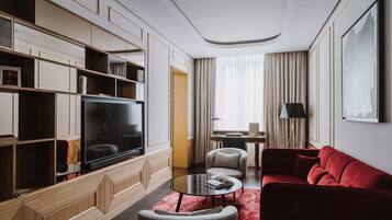 Una habitación de hotel moderna con una televisión de pantalla plana, un sofá rojo, una mesita de centro redonda, una estantería y un escritorio con lámpara.