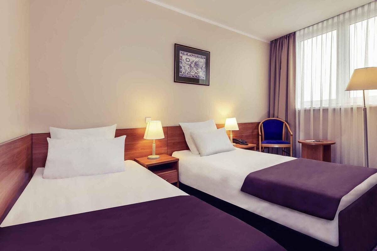 A hotel room with two beds, a nightstand with a lamp, a chair, and a small table.