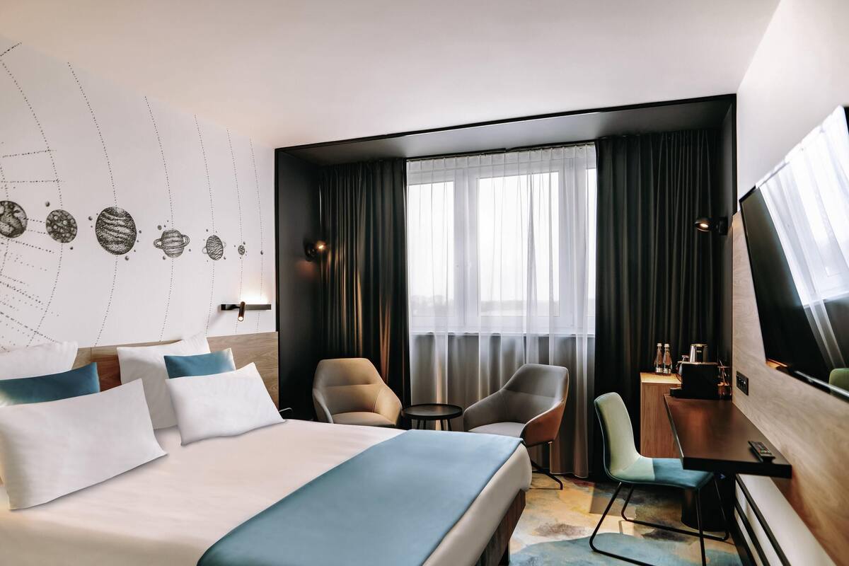 A hotel room with a large bed, two armchairs, a desk, a TV, and a window with curtains.