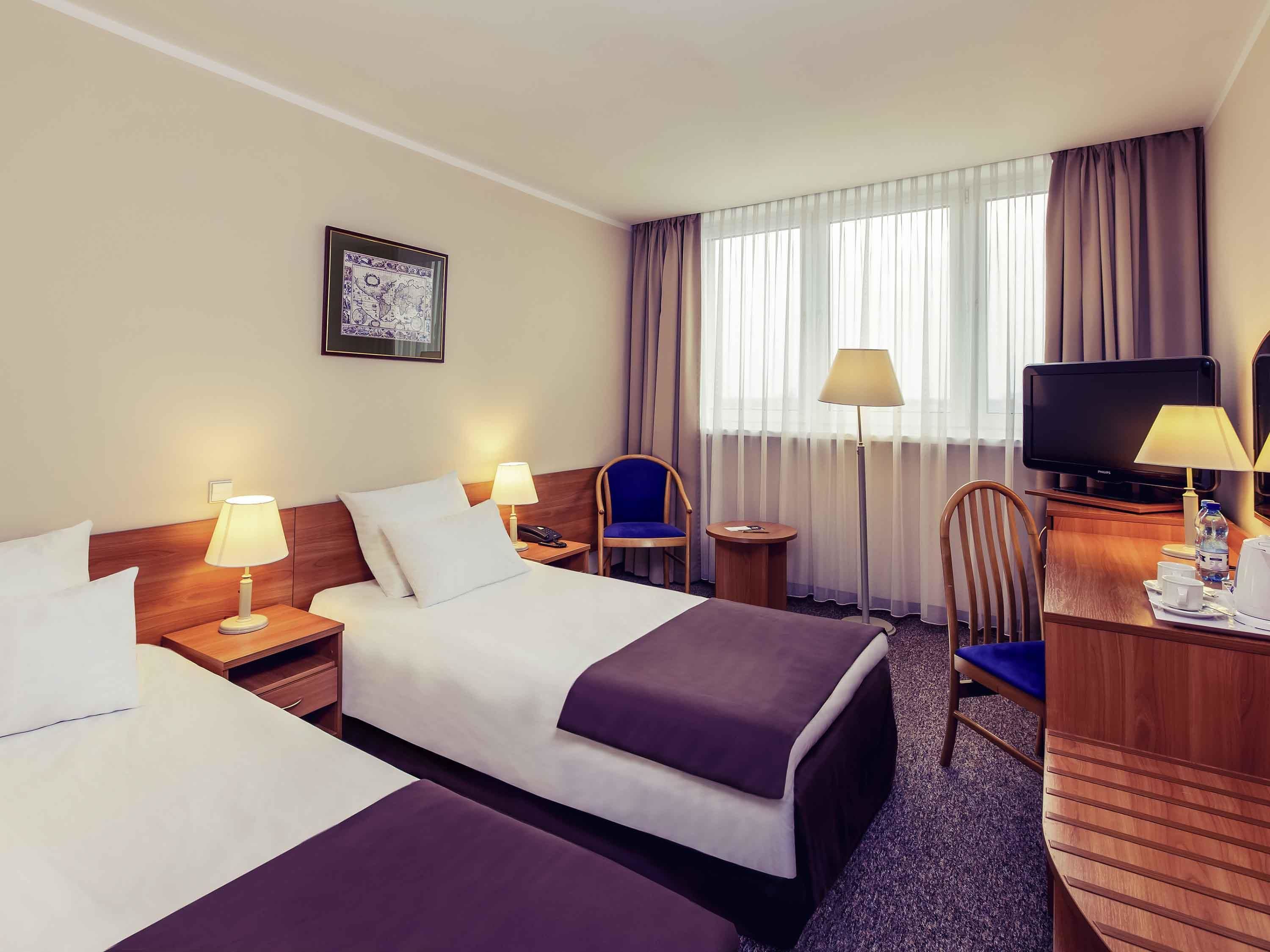 Ein Hotelzimmer mit zwei Betten, einem Schreibtisch, einem Fernseher, einem Sessel und einem Fenster mit Vorhängen.