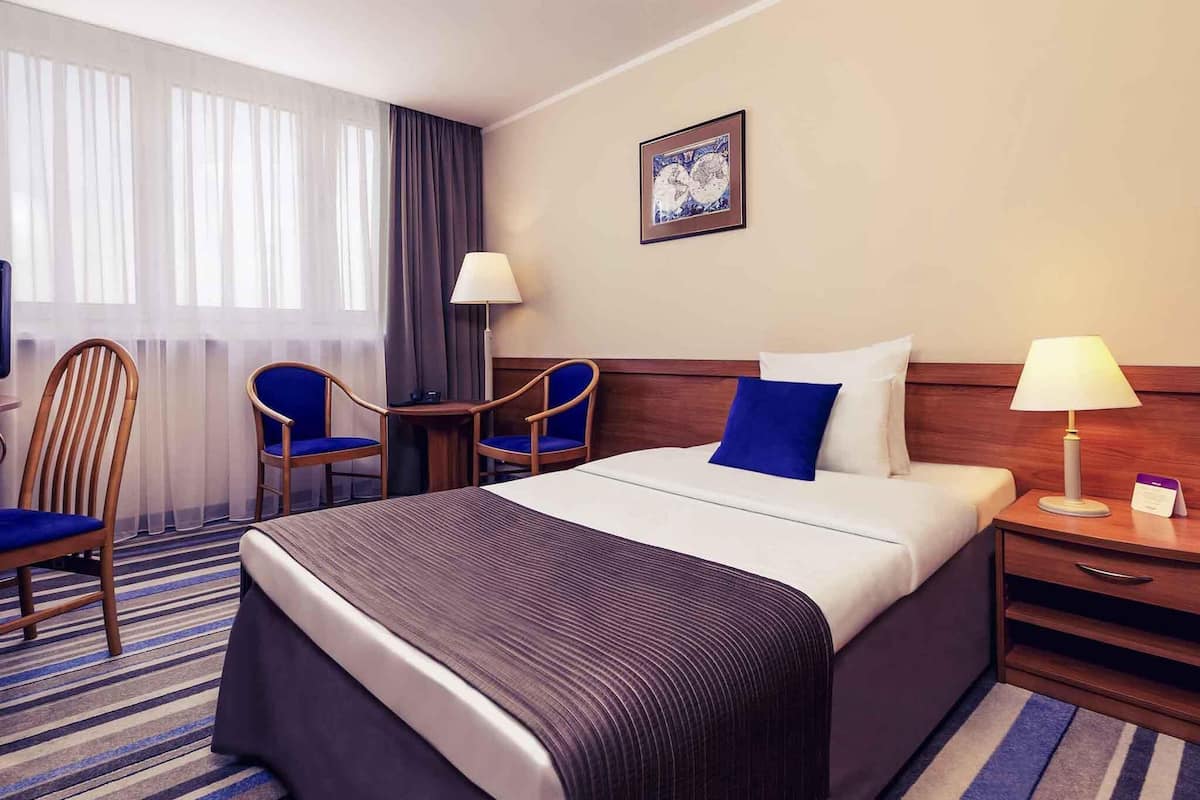 A hotel room with a large bed, two chairs, a small table, a nightstand with a lamp, and a painting on the wall.