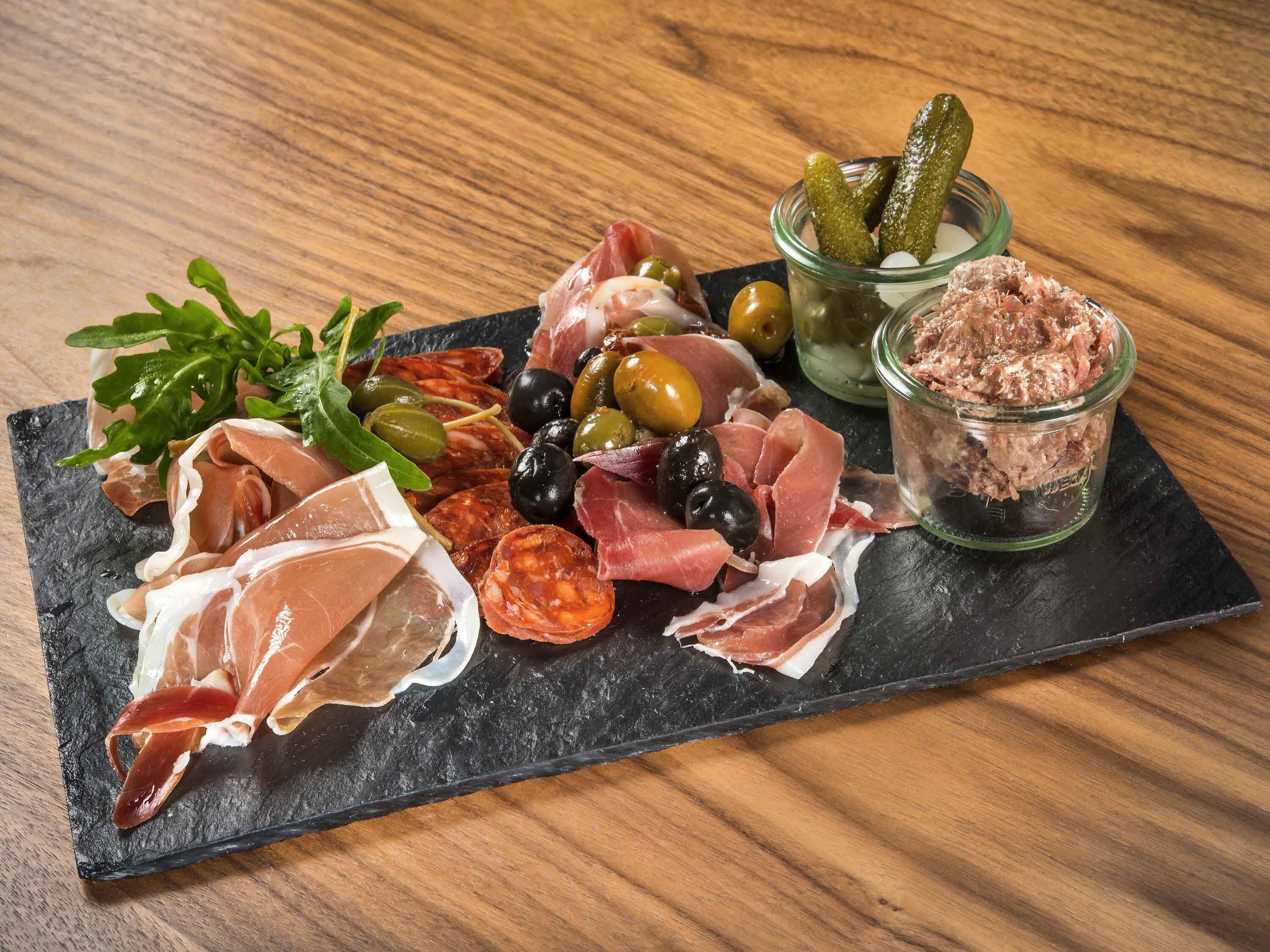 Une planche de charcuterie comprenant différentes sortes de viandes, des olives, des cornichons et des légumes verts, le tout présenté sur une ardoise.
