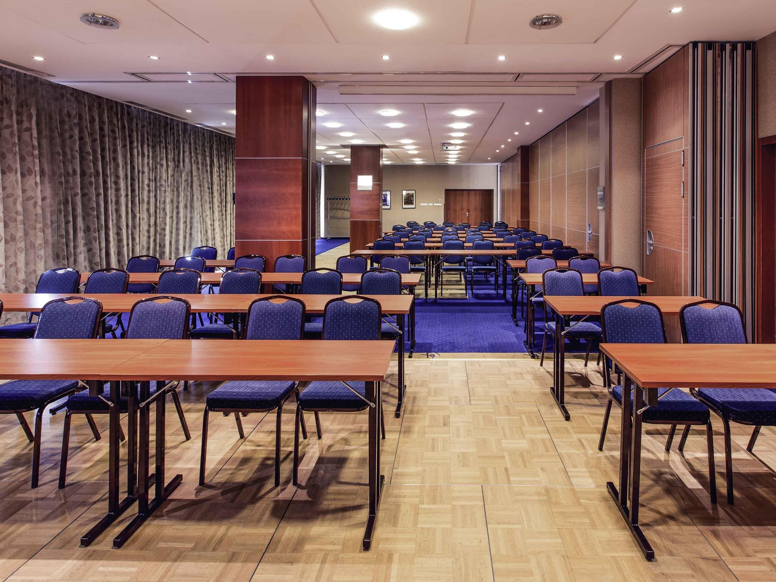 A conference room with rows of tables and chairs, wooden flooring, and a patterned wall.