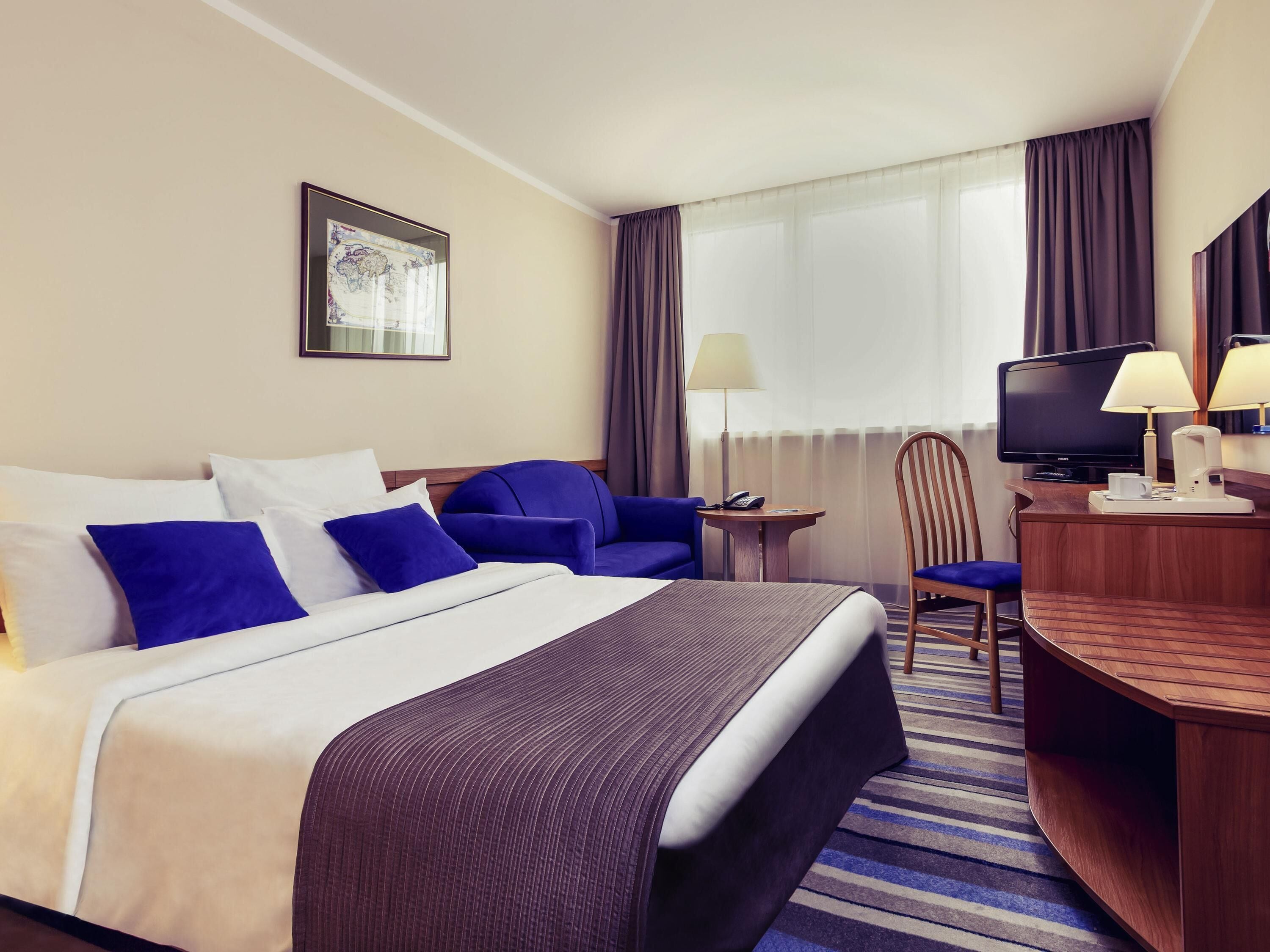 A hotel room with a bed, a desk with a television, a chair, and a lamp.