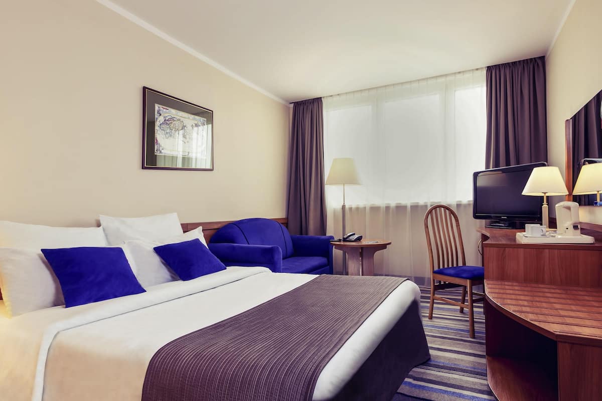 A hotel room with a bed, a desk with a television, a chair, and a lamp.