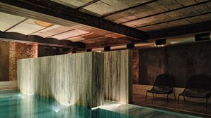Uma piscina interior moderna com divisória de madeira, duas cadeiras de vime e teto de concreto com vigas aparentes.