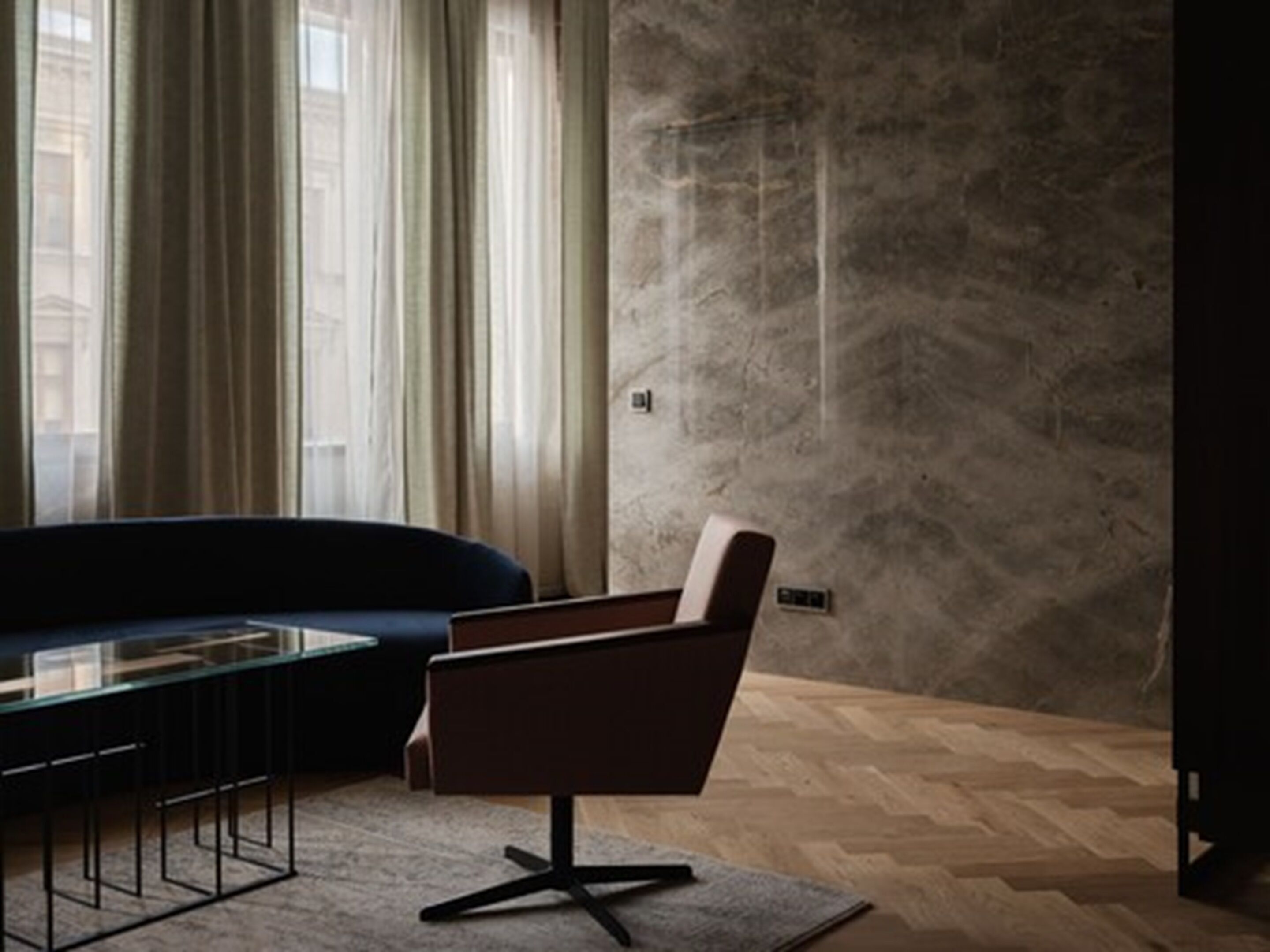 A modern living room with a dark blue sofa, a brown armchair, and a glass coffee table. The room has a herringbone wood floor and a textured stone wall.