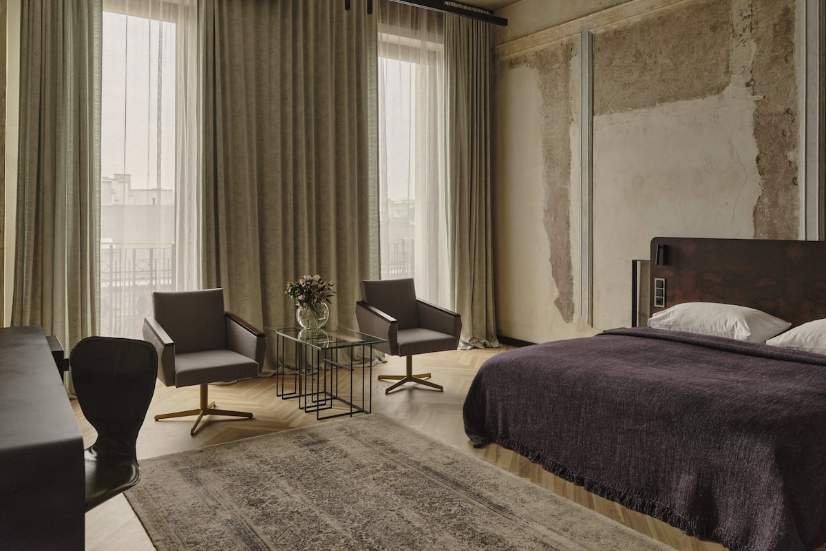Habitación de hotel con cama, dos sillones, una mesita con un jarrón y vistas a la ciudad a través de amplias ventanas.