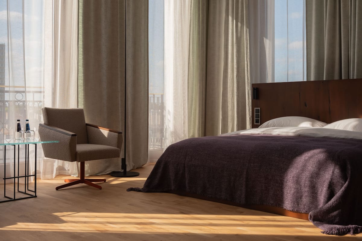 Habitación de hotel con cama, una silla, una mesita de cristal con botellas y cortinas de encaje.