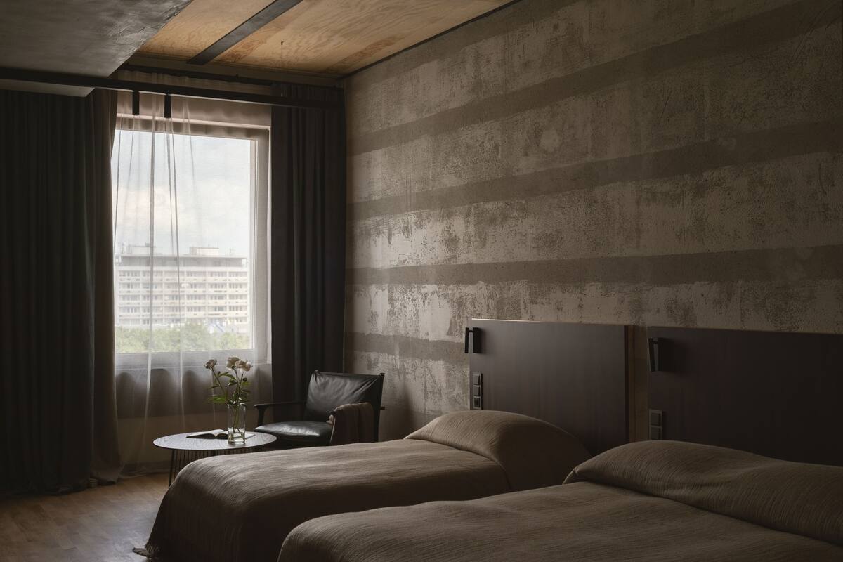 Habitación de hotel con dos camas, una mesita con un jarrón de flores, una silla y vistas a un paisaje urbano a través de una ventana con cortinas.