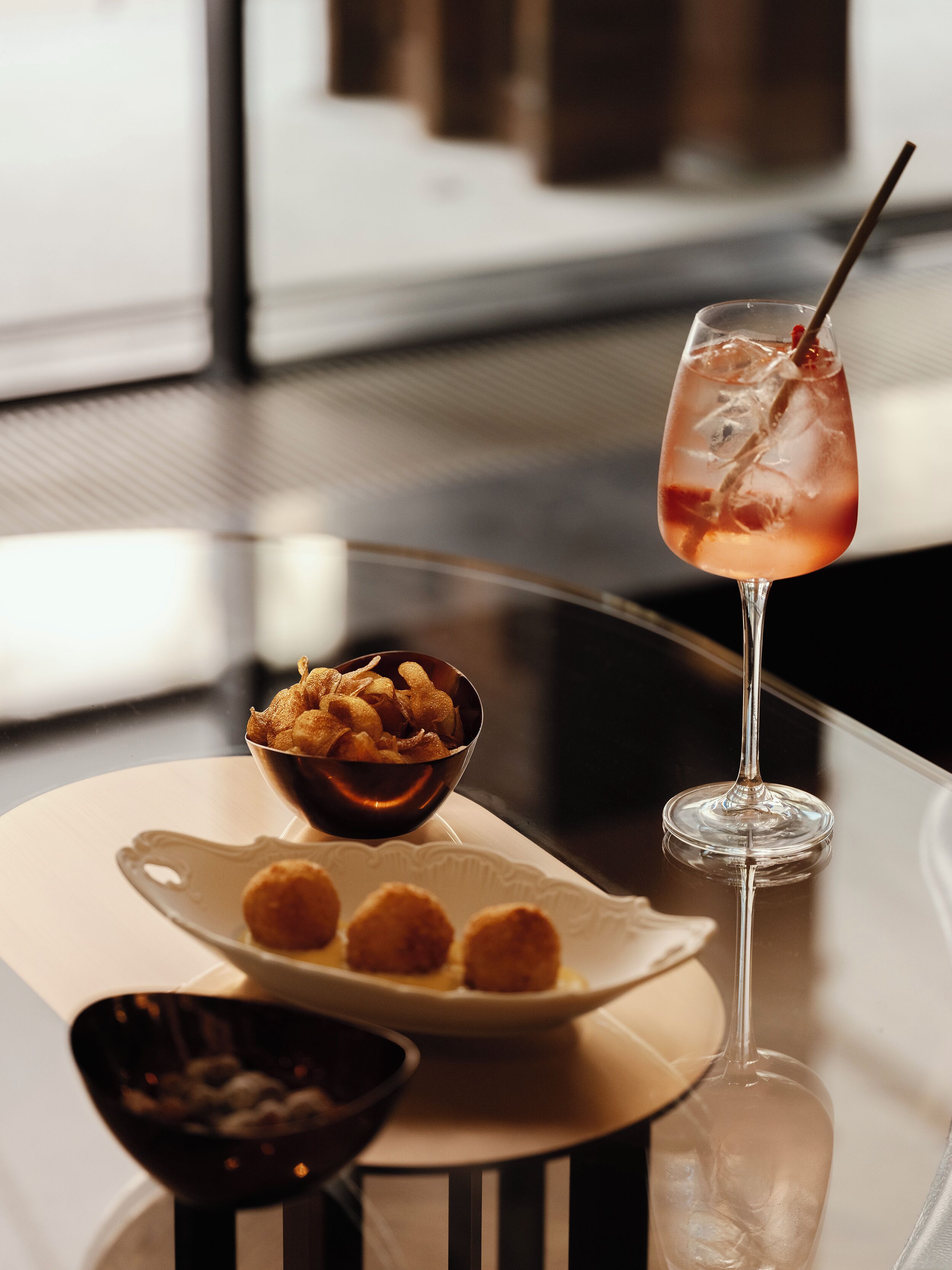 A table with a glass of a pinkish drink, a bowl of fried snacks, and a plate with round items.