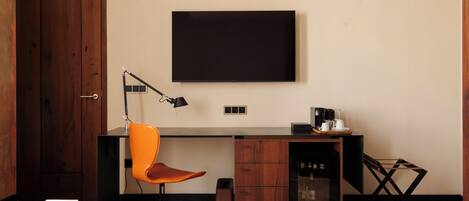 Un espacio de oficina moderno con un escritorio de madera, una silla naranja, una lámpara de escritorio negra, una máquina de café y un televisor fijado a la pared.