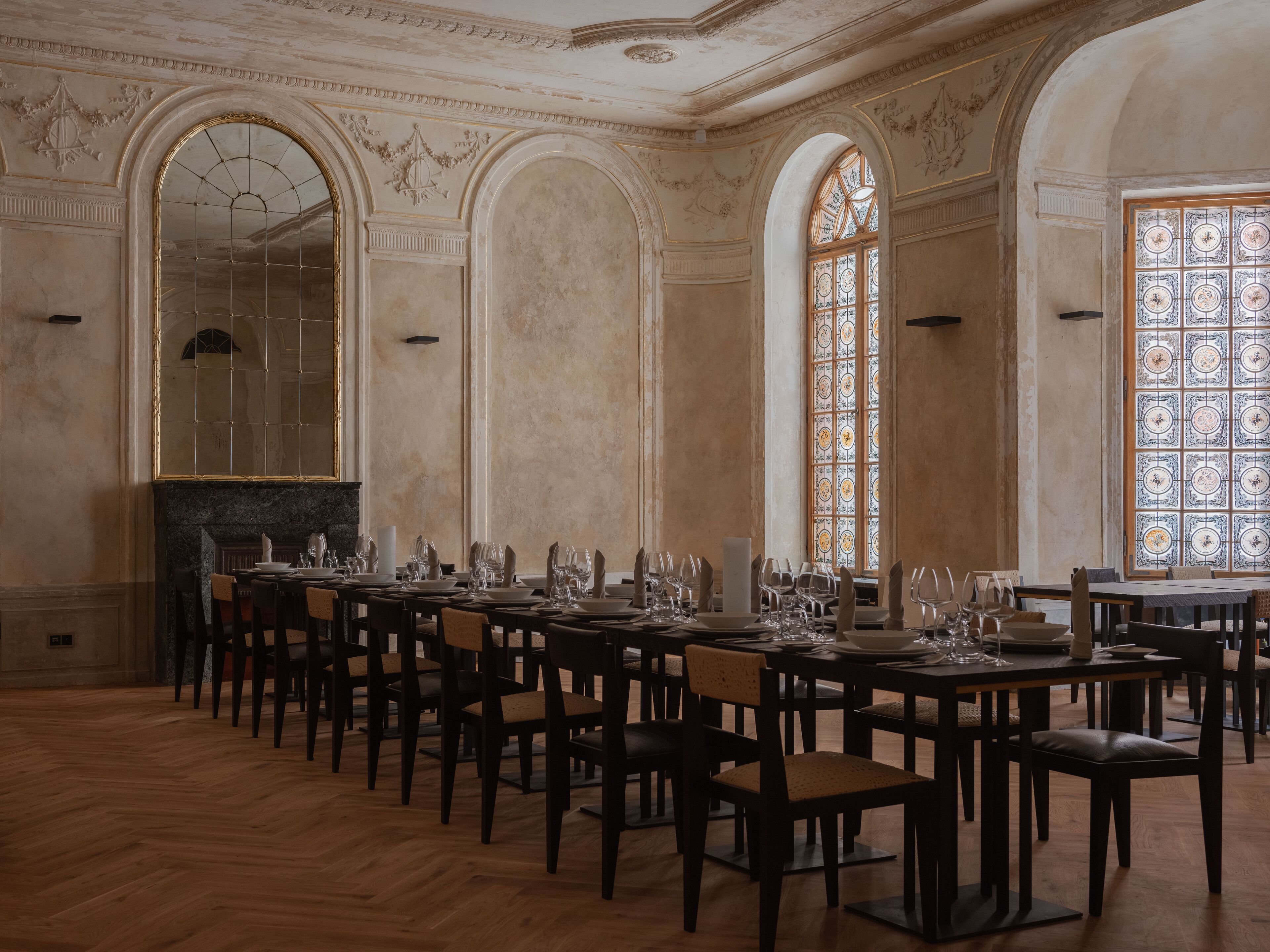 A formal dining room with multiple tables set for a meal, high ceilings with decorative moldings, and large windows with stained glass.