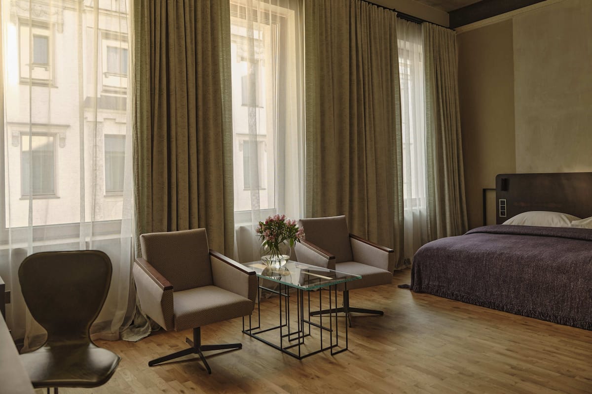 Habitación de hotel con cama, dos sillones, una mesa de centro de vidrio y vistas a edificios a través de cortinas translúcidas.