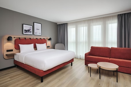A hotel room with a bed, a red sofa, two small tables, and a window with curtains.