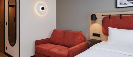 Ein Hotelzimmer mit einem Bett, einer roten Couch, einem Nachttisch, einer Wandlampe und einem gerahmten Bild an der Wand.