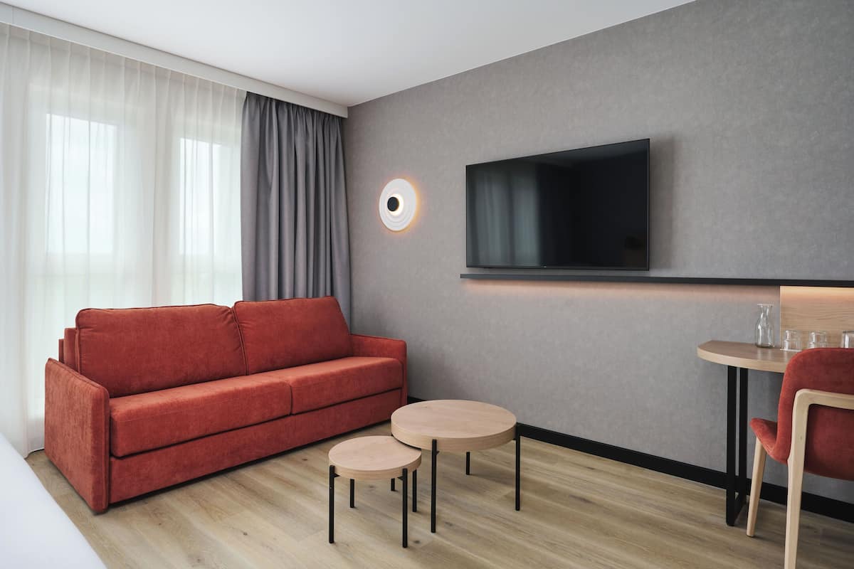 A modern hotel room with a red sofa, two small round tables, a TV mounted on the wall, and a large window with sheer curtains.