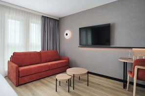Una habitación de hotel moderna con un sofá rojo, dos mesas redondas pequeñas, un televisor montado en la pared y un amplio ventanal con cortinas de gasa.