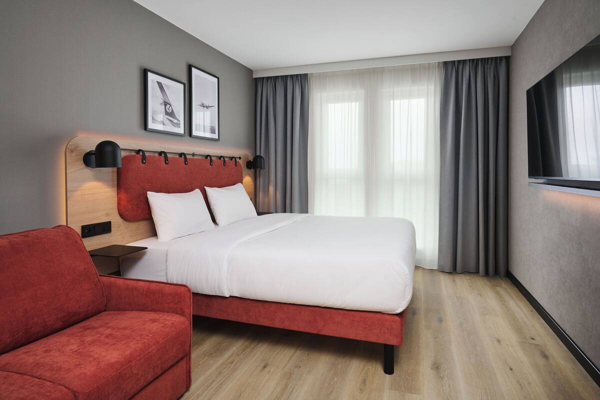 A modern hotel room with a bed, a red armchair, a wooden headboard, and two framed pictures on the wall.