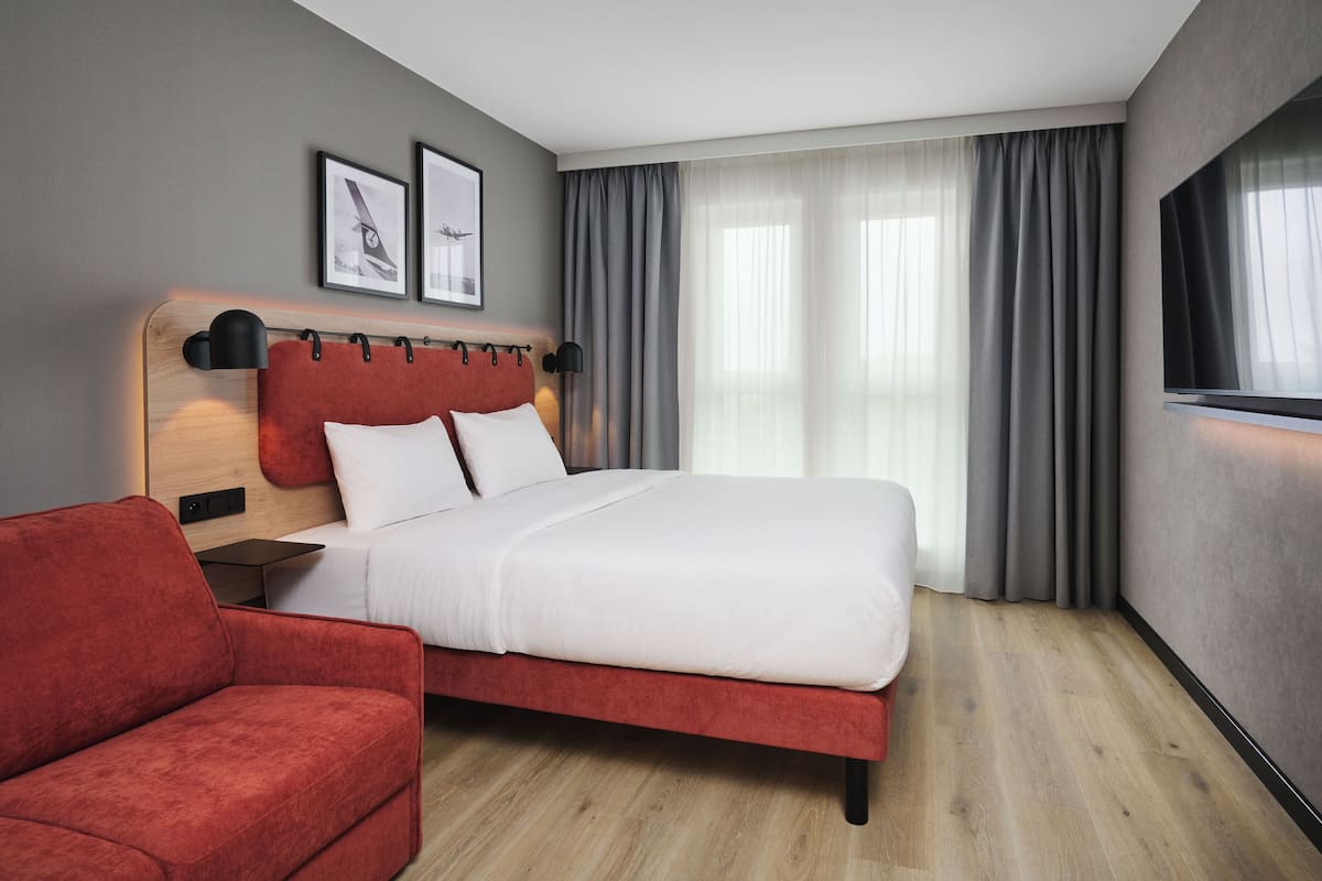 Quarto de hotel moderno com uma cama, uma poltrona vermelha, uma cabeceira de madeira e duas imagens emolduradas na parede.