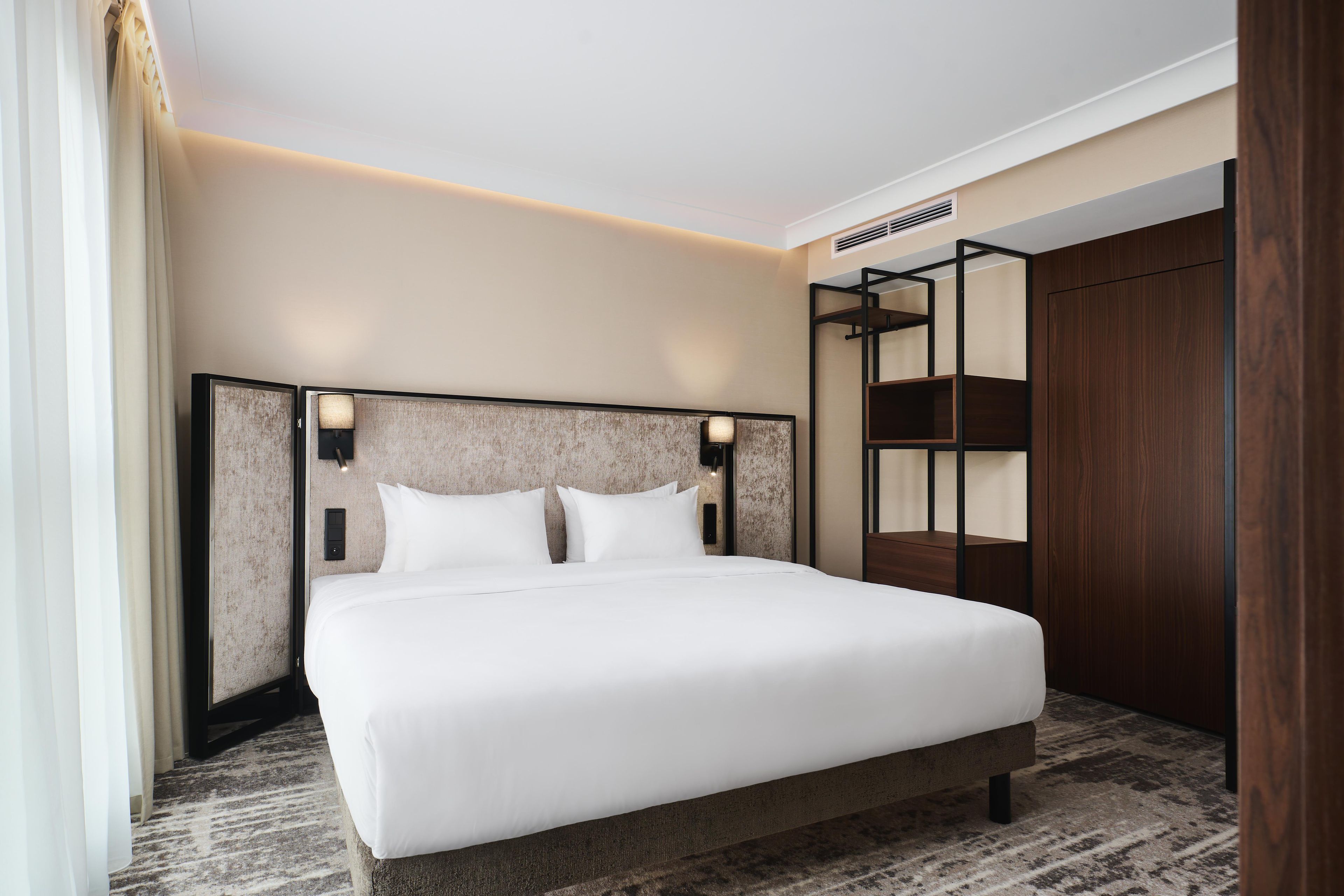 A modern hotel room with a large bed, a headboard with integrated lighting, a built-in wardrobe, and a carpeted floor.