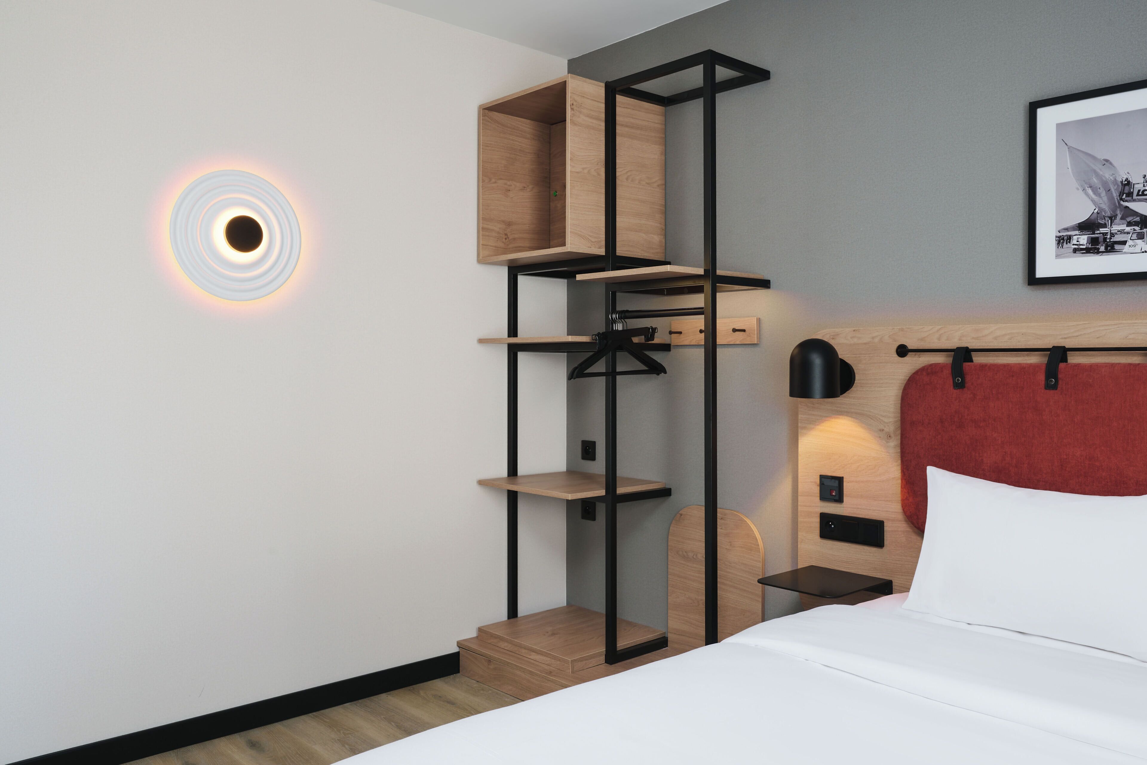 Une chambre d’hôtel moderne avec un lit, une tête de lit rouge, une étagère fixée au mur et un luminaire circulaire.