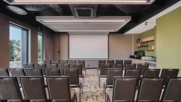 A conference room with rows of chairs facing a projection screen, a coffee station, and a view of the outdoors through large windows.