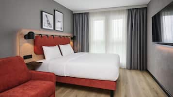 A hotel room with a bed, a red armchair, a bedside table, a wall-mounted TV, and two framed pictures.
