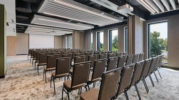 Un salón de conferencias con filas de sillas, un proyector y ventanas amplias.