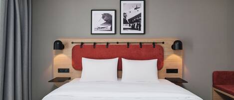 Une chambre d’hôtel avec un lit, deux tableaux encadrés au mur, une tête de lit rouge et une chaise rouge.
