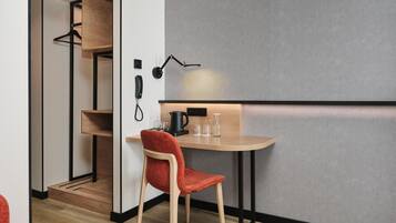 A compact hotel room with a wooden desk, a red chair, a black desk lamp, and a telephone.
