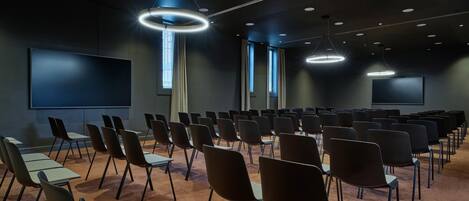 Een vergaderruimte met rijen stoelen, een groot scherm en plafondlampen.
