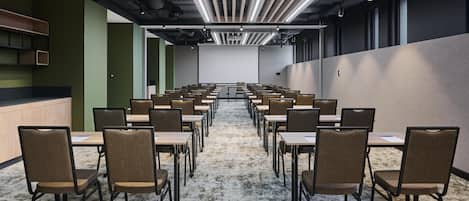 A conference room with rows of tables and chairs, a projector screen, and a ceiling with linear lighting fixtures.