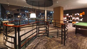 Um espaço interno moderno com mesa de bilhar, estantes de livros e vista panorâmica da cidade através de grandes janelas.