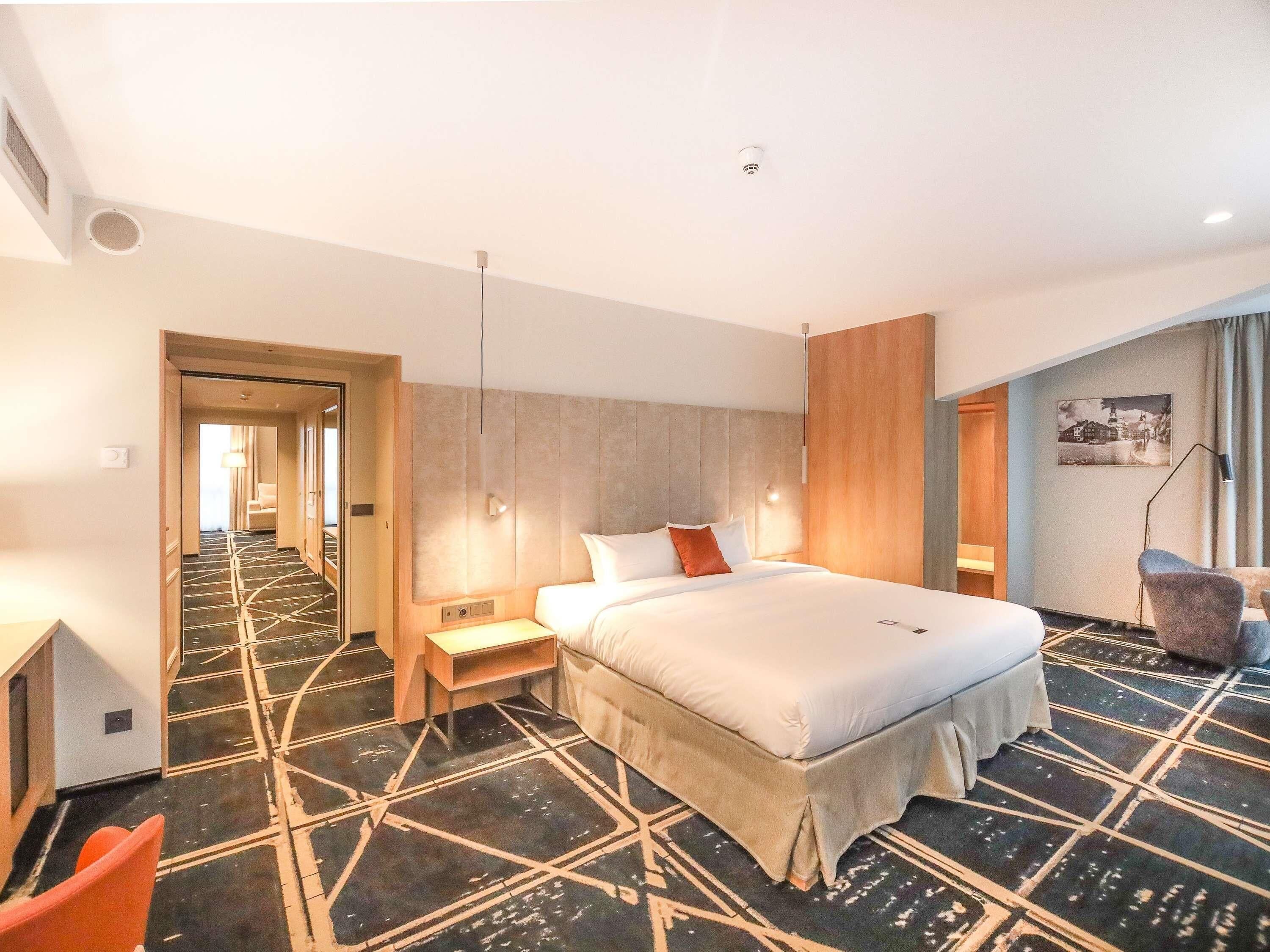 A hotel room with a bed, a desk, a chair, a lamp, and a picture on the wall.